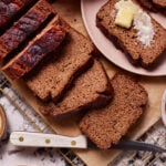 Several slices of banana bread on a cutting board, with one slice on a pink plate topped with a pat of butter. A serrated knife with a white handle is placed next to the board. The scene is set on a marble surface with a cooling rack underneath.