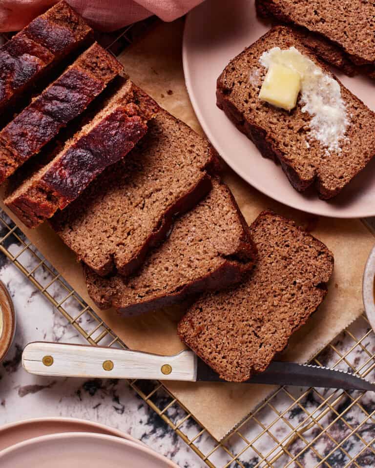 Several slices of banana bread on a cutting board, with one slice on a pink plate topped with a pat of butter. A serrated knife with a white handle is placed next to the board. The scene is set on a marble surface with a cooling rack underneath.