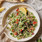 A bowl of the grain free tabouleh salad, topped with a lemon wedge. A fork sits in the bowl, and a cutting board with lemons and salt can be seen in the background
