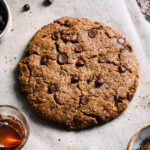 A giant chocolate chip cookie laying on a sheet of parchment paper. Chocolate chips can be seen scattered across the parchment paper