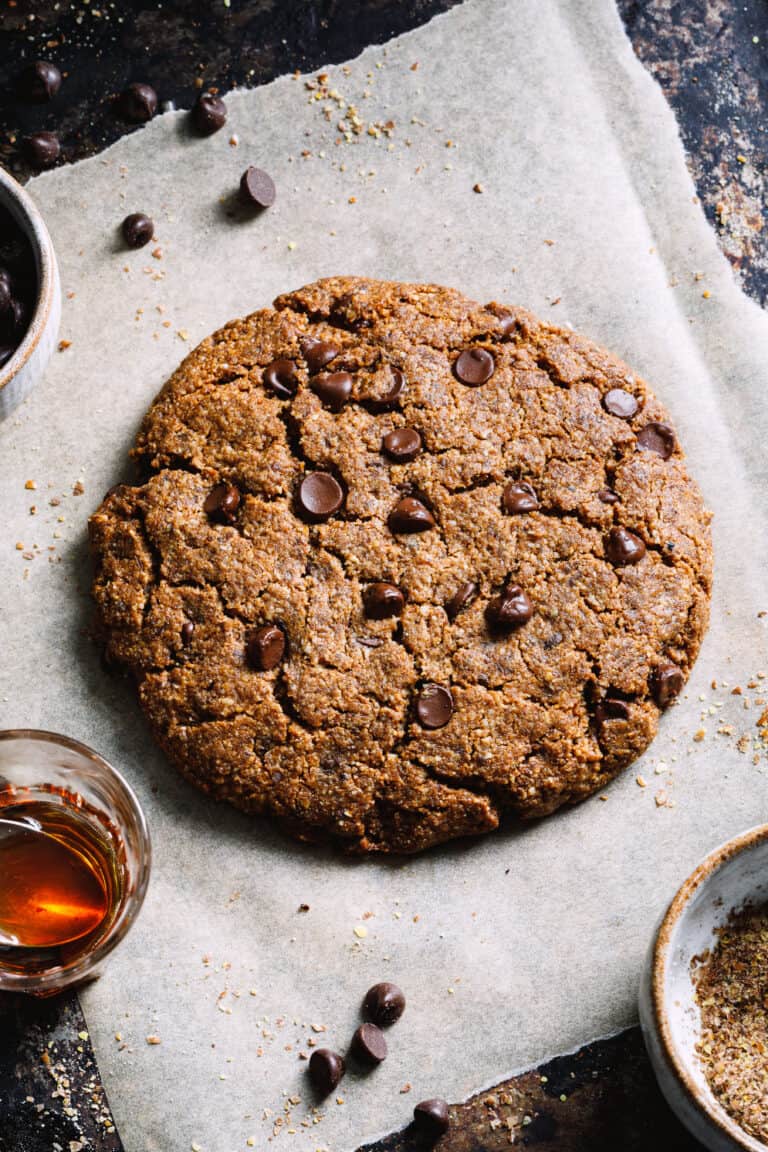 A giant chocolate chip cookie laying on a sheet of parchment paper. Chocolate chips can be seen scattered across the parchment paper