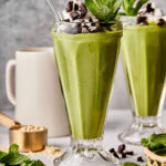A tall glass of the healthy, dairy-free shamrock shake topped with whipped cream, crushed Oreos, mint leaves, and a glass straw. A mug is in the background with ingredients scattered on the ground