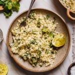 A large plate of the Copycat Chipotle Cauliflower Cilantro Lime Rice, with a spoon dug into it. A larger pot of rice sits off to the right corner, and a plate of cilantro and lime sits off to the left.