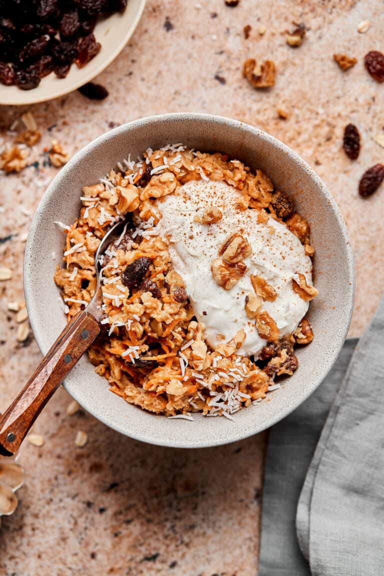 A bowl of the carrot cake cauliflower oats, on a countertop, with ingredients scattered beside it.