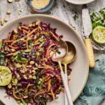 A bowl of the cabbage carrot salad with tangy peanut dressing, on a cutting board with ingredients scattered around it. A long serving spoon sits in the bowl.