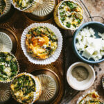 A plate of the spinach feta egg muffins, lined up on top of each other. A small bowl of feta cheese rests to the side