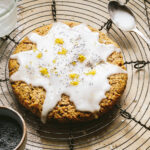 A Giant lemon poppy seed cookie, laying on wire cooling rack. A spoon and a small bowl and be seen in the background.