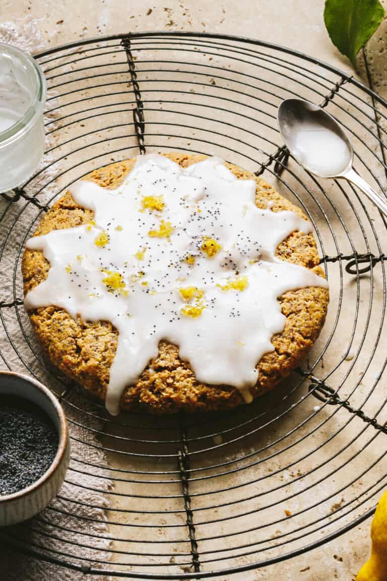 A Giant lemon poppy seed cookie, laying on wire cooling rack. A spoon and a small bowl and be seen in the background.