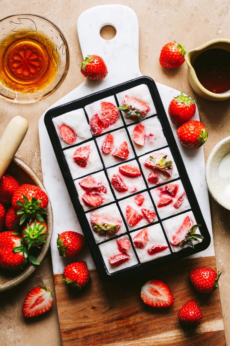 an ice cube tray full of the high protein strawberry yogurt cubes, sitting on top of a marble cheeseboard. A basket of strawberries and a small bowl of maple syrup can be seen off to the side