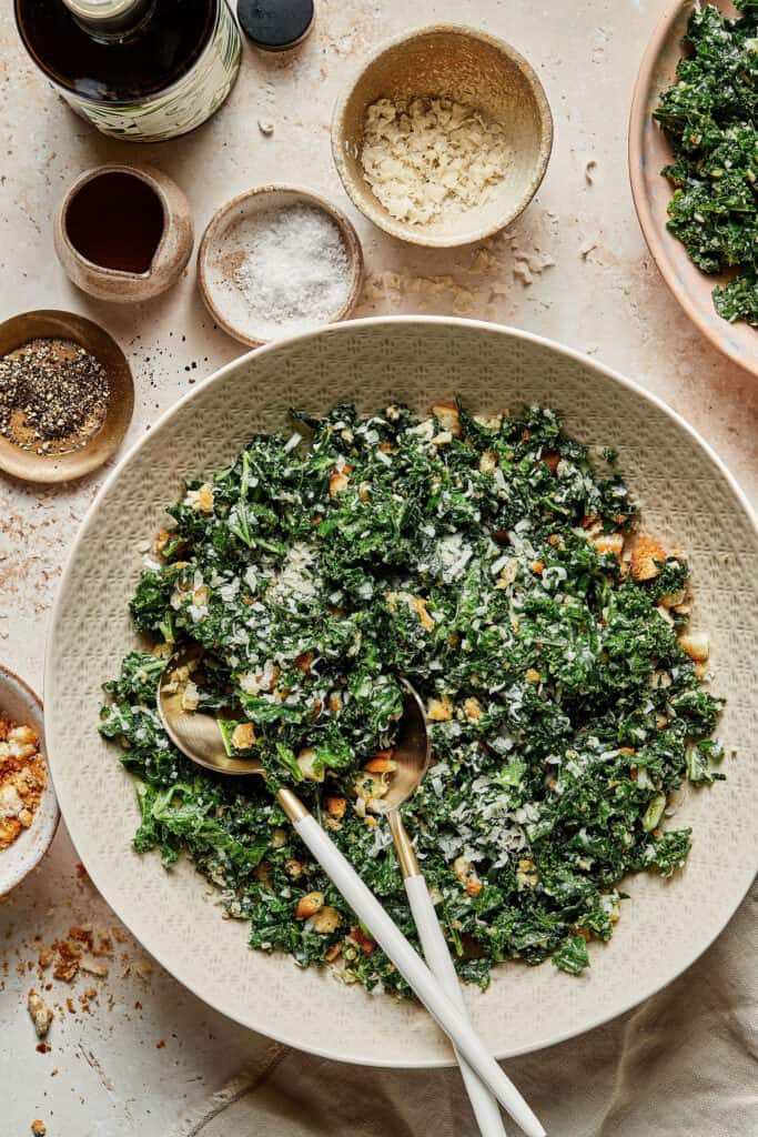 A large bowl of the lemony garlic parmesan kale salad, with two forks sticking out of the bowl. Smaller bowls with the separate ingredients can be seen in the background.