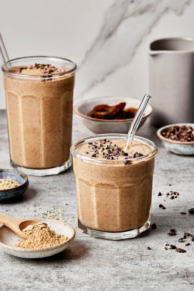 Two glasses of the copycat Erewhon Maca bomb smoothie, with one in the foreground and one in the background. Different bowls of ingredients can be seen further in the background.