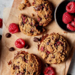 Three of the paleo raspberry chocolate chip cookies, laid out on a sheet of parchment paper. A small bowl of raspberries can be seen off to the side.