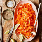 A large bowl of shredded carrots, sitting on top of a cutting board alongside smaller bowls filled with the other ingredients needed to make this salad