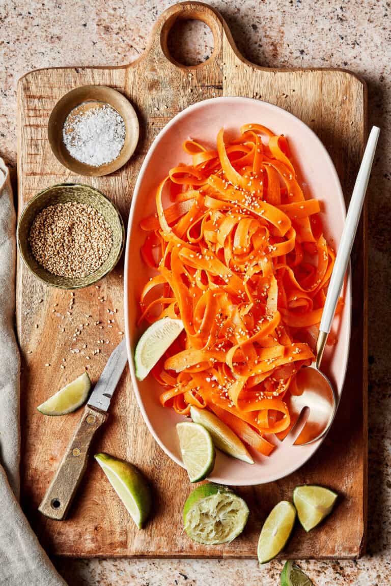 A large bowl of shredded carrots, sitting on top of a cutting board alongside smaller bowls filled with the other ingredients needed to make this salad