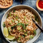 A bowl of rice noodles topped with chopped peanuts, green onions, and mint leaves, garnished with lime wedges. A small bowl of sesame seeds, chopped peanuts, and a dollop of red sauce are nearby, all set on a blue textured surface.