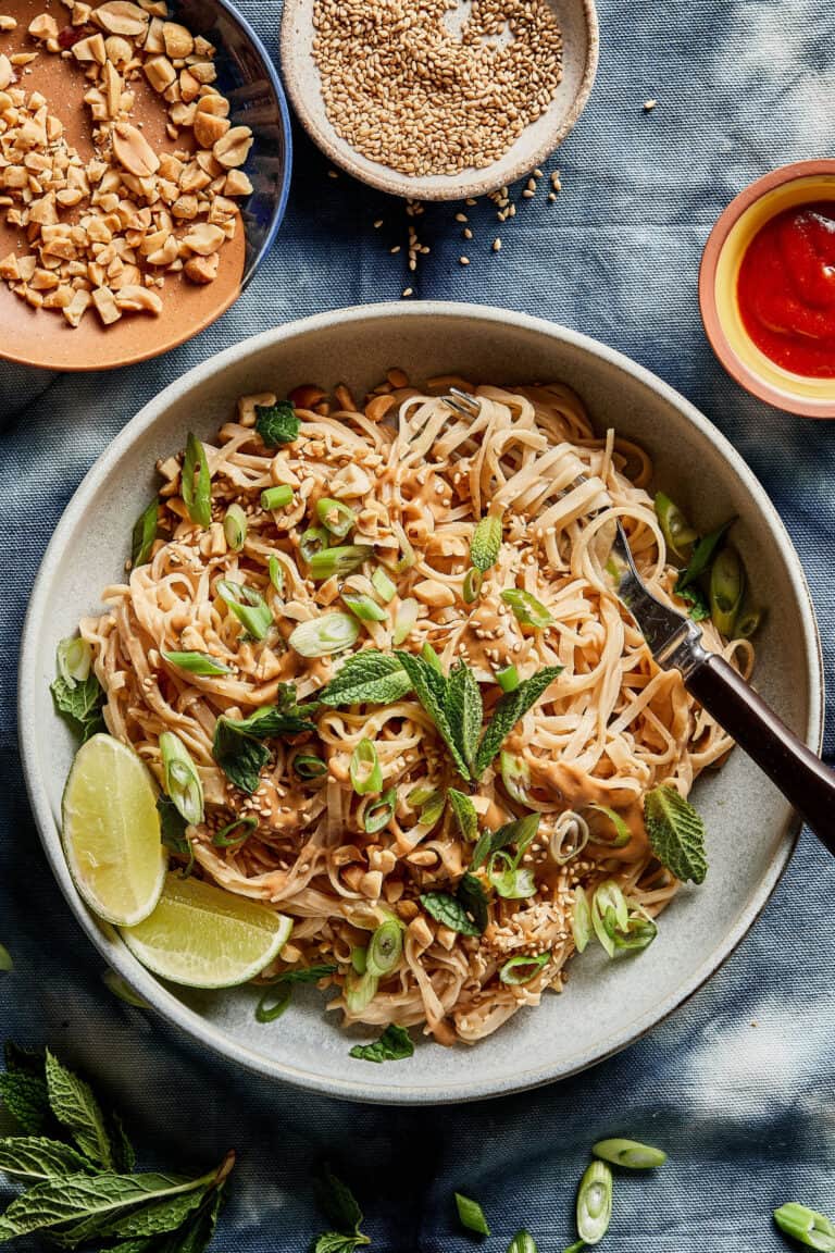 A bowl of rice noodles topped with chopped peanuts, green onions, and mint leaves, garnished with lime wedges. A small bowl of sesame seeds, chopped peanuts, and a dollop of red sauce are nearby, all set on a blue textured surface.