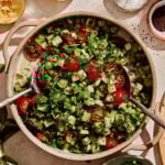 A large bowl of the Armenian cucumber tomato salad, on a countertop surrounded by different ingredients