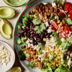 A large bowl of the chicken taco salad, on a countertop, surrounded by a small bowl of dressing, a sliced avocado, a plate of shredded chicken, and lime and cilantro.