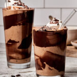 Two glasses of the chocolate cookie milkshake smoothie, in tall glasses topped with whipped cream.
