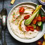 A bowl of the healthy white bean dip, topped with chopped cucumbers and tomatoes. A spoon can be seen resting next to the bowl.