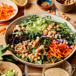 A large bowl of the moroccan inspired chickpea carrot salad, on a countertop surrounded by the different salad ingredients.