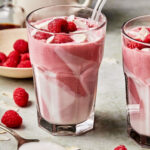 A glass of the raspberry coconut cloud smoothie, topped with fresh raspberries and a straw. A second glass sits to the side of it, and a bowl of raspberries rests behind it.