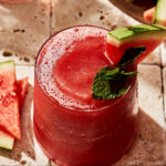 A tall glass of the watermelon frosé, with a watermelon slice and a sprig of fresh mint