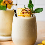 A tall glass of the 3-ingredient high protein dole whip, sitting on a counter with a slice of pineapple and a sprig of fresh mint in the glass