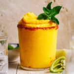 A tall glass of the frozen mezcal mango margarita, on a countertop with a lime wedge and jalapeño slices