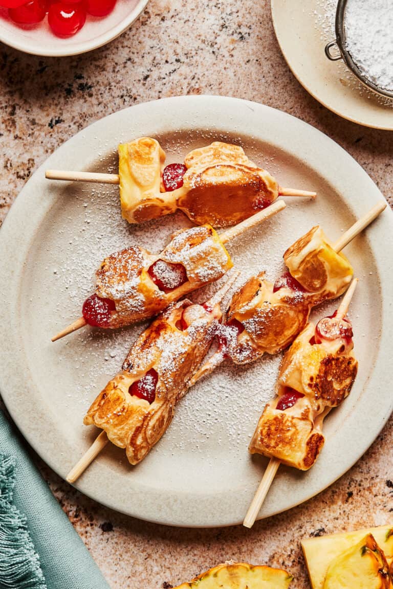 A plate of the pineapple upside down pancake dippers, sitting on a countertop surrounded by bowls of fresh pineapple and maraschino cherries