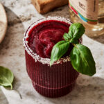A glass of the basil mixed berry mezcal margarita, topped with a sprig of basil and sitting on a countertop.