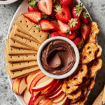 A platter of the healthy brownie batter sugar cookie dip, alongside sliced apples, strawberries, graham crackers, and pretzels.