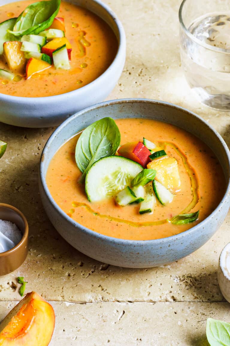 A bowl of the Summer Peach Gazpacho, sitting on a countertop. Another bowl of the peach gazpacho sits behind it.