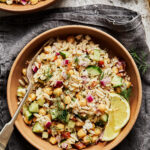 A bowl of the chickpea orzo salad, witting on a napkin on a table. A second plate of salad and a fork can be seen in the background.
