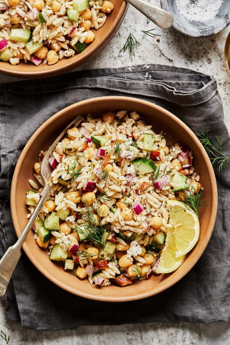 A bowl of the chickpea orzo salad, witting on a napkin on a table. A second plate of salad and a fork can be seen in the background.