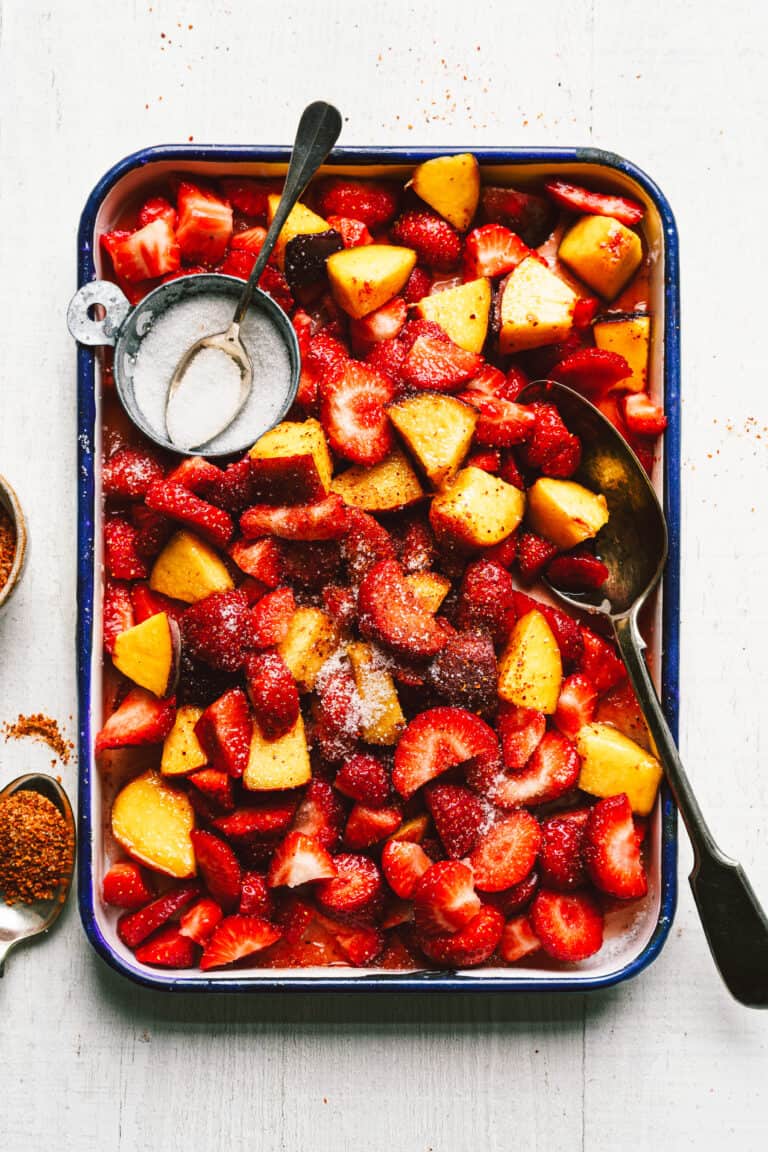 A tray of the sweet & sour tajin peaches & strawberries, sitting on a countertop. A bowl of sugar sits nestled into the peaches and strawberries