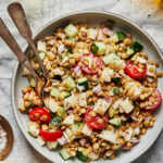 A bowl of fresh salad contains lentils, cucumber, cherry tomatoes, red onions, and small cheese cubes. A fork and spoon rest in the bowl. Surrounding the salad are small dishes and a glass, presumably containing dressing and seasonings.