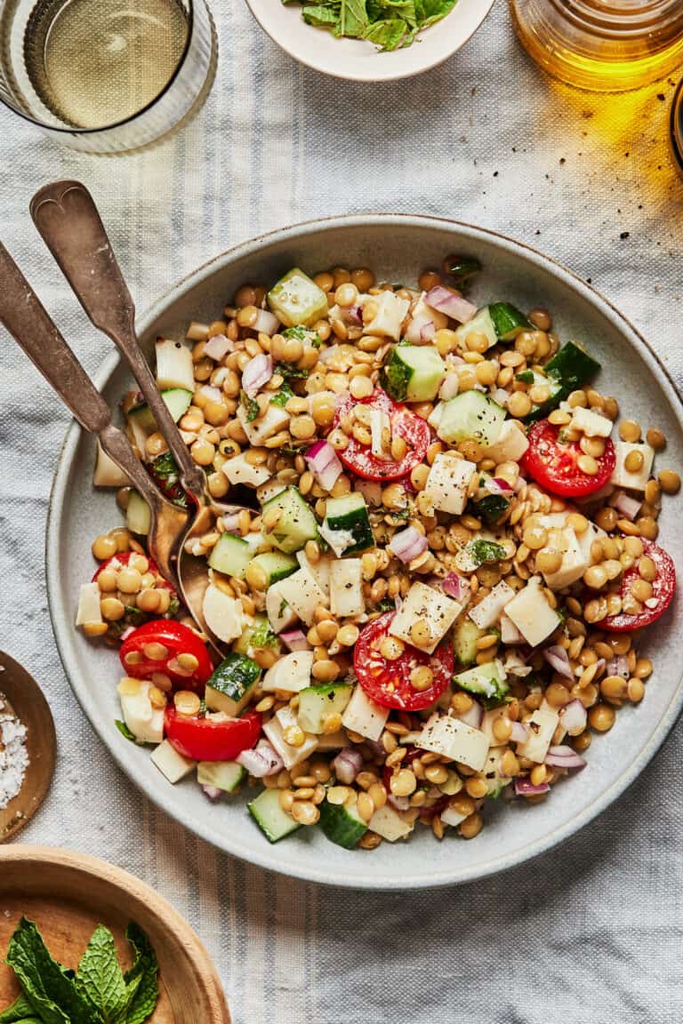 A bowl of fresh salad contains lentils, cucumber, cherry tomatoes, red onions, and small cheese cubes. A fork and spoon rest in the bowl. Surrounding the salad are small dishes and a glass, presumably containing dressing and seasonings.