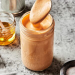 A jar of the creamy chipotle dressing, sitting on a countertop, with a spoon drizzling some of the dressing back into the jar. A few of the ingredients needed to make this dressing can be seen in the background.
