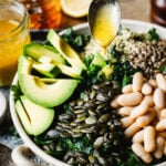 A large bowl of the copycat Erewhon kale and white bean salad, sitting on a countertop, with the dressing being actively poured over it