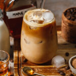 A tall glass of the copycat Starbucks pumpkin cream cold brew, sitting on a cutting board on a countertop. Various ingredients can be seen in the background.