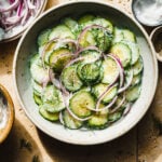 A bowl of the no mayo creamy cucumber salad, sitting on a countertop alongside different small bowls of ingredients.