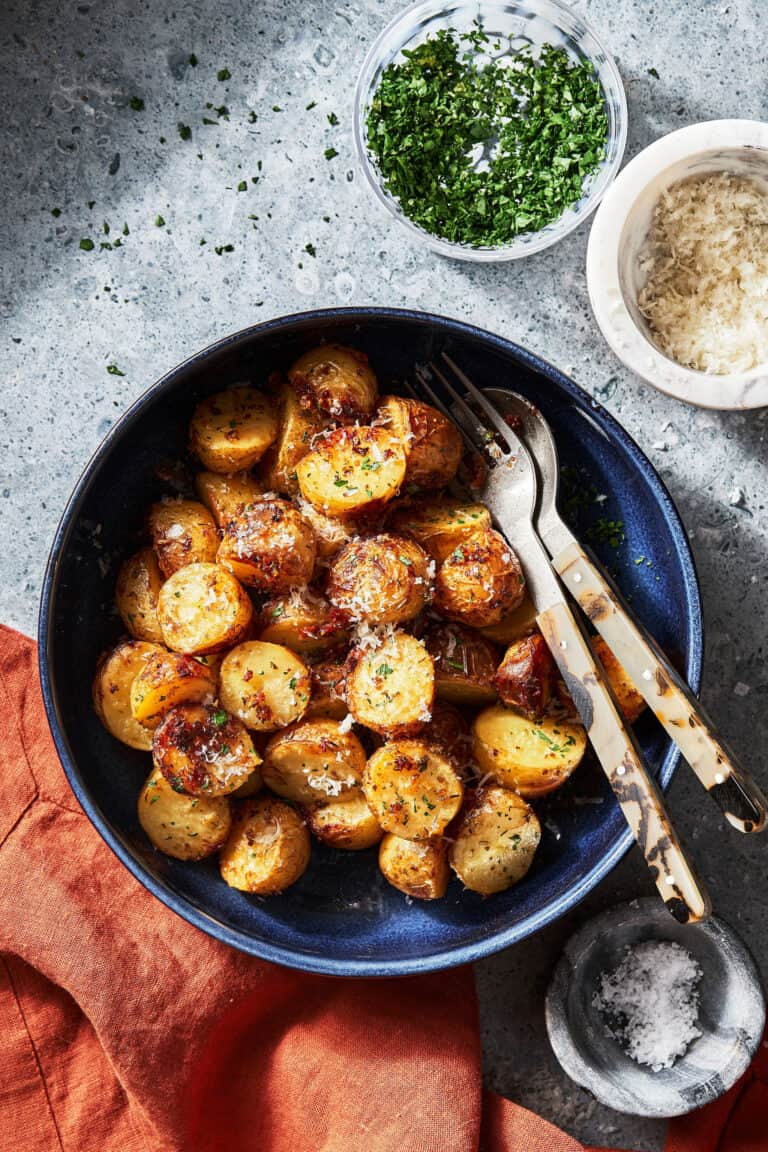 A bowl of the viral tiktok crispy parmesan potatoes, sitting on a countertop surrounded by bowls of parmesan and parsley
