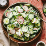A big bowl of the creamy cucumber radish salad, sitting on a countertop surrounded by various ingredients