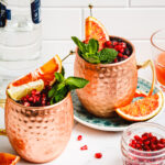 Two glasses of the pomegranate Moscow mules, sitting on a countertop. A small jar of pomegranate seeds and a bottle of vodka can be seen in the background