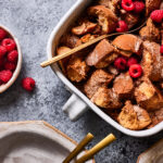 A platter of the lightened up French toast casserole, sitting on a countertop, alongside a plate, knife, and a bowl of berries