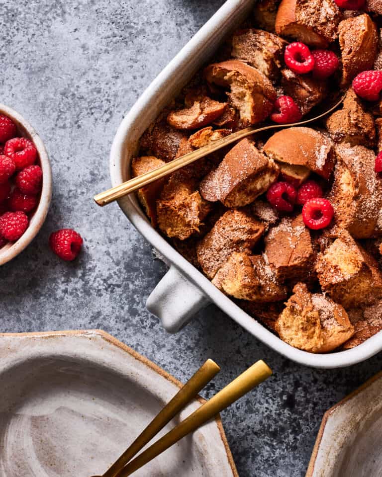 A platter of the lightened up French toast casserole, sitting on a countertop, alongside a plate, knife, and a bowl of berries