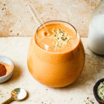 A glass of the sweet potato apple pie smoothie, topped with hemp seeds and with a straw stuck into it. A few of the ingredients needed to make this smoothie can be seen off to the side