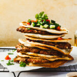 A stack of the meat stuffed pitas (arayes), lined up on a wire cooling rack and topped with chopped tomato, cucumber, and fresh parsley