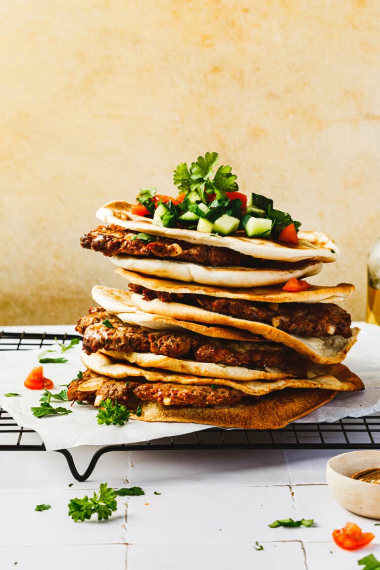 A stack of the meat stuffed pitas (arayes), lined up on a wire cooling rack and topped with chopped tomato, cucumber, and fresh parsley